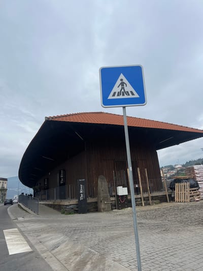 This image depicts a pedestrian crossing sign and a distinctive building in Peso da Régua, Portugal, under an overcast sky.

In the foreground, a blue square pedestrian crossing sign with a white triangular inset containing a black walking figure is prominently positioned on a silver pole, slightly right of center. Below and to the left of the sign, the edge of an asphalt road with a partial white pedestrian crossing strip is visible, alongside a grey cobblestone sidewalk that extends towards the midground.

The primary structure in the midground is a two-story building with dark brown vertical wooden siding and a wide, curved terracotta-tiled roof with a significant overhang, particularly noticeable on the left. Multiple black signs bearing the text "The River" and "RESTAURANT" are affixed to the building's facade and a black metal railing that runs along its front. To the right of the building's entrance, a large, rough-hewn stone column stands next to a modern white electrical box mounted on the wall. Further to the right, a pile of wooden pallets and what appears to be construction materials (possibly red bricks or bags covered by a black tarp) are stacked on a patch of uneven ground.

In the background, a road curves towards the left, with a white sedan and a dark SUV parked along its edge. Distant hillsides, covered with residential buildings and vegetation, rise under the grey, cloudy sky. No individuals are clearly visible or engaged in specific actions within the frame.
