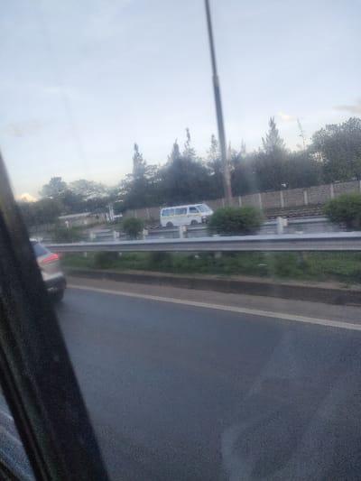 The image depicts a highway scene in Murera ward, Kenya, viewed from inside a moving vehicle. The immediate foreground on the left shows a dark, blurred portion of the vehicle's interior. A paved asphalt road with a solid white line is visible in the lower-left to mid-right. A light-colored vehicle, possibly silver or grey, is partially visible from the rear on the far left lane. A silver-colored metal guardrail spans horizontally across the mid-ground, separating the primary road from a grassy median containing sparse green shrubs. Beyond the median, a white van with yellow horizontal stripes on its side is visible, facing away from the camera's direction, positioned on an adjacent road section or lane. A tall, dark utility pole extends vertically from the mid-ground. The background features a low, light-colored stone wall running parallel to the road, backed by a dense line of various trees, some conical. The sky overhead is overcast and light grey. No individuals are visible.
