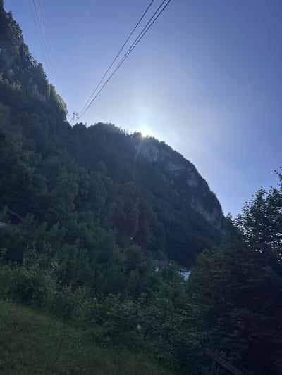 A wide-angle, low-angle shot captures a steep, tree-covered mountainside under a clear blue sky. The sun shines brightly, partially obscured by the mountain, creating a lens flare. Four parallel cable car lines ascend diagonally from the lower left to the upper right of the frame, leading to an unseen station. One cable car structure is visible mid-way up the cables. The mountain is densely forested, with some rocky outcrops visible near the summit on the right side. In the lower left quadrant, a small wooden building is nestled amongst the trees. The foreground consists of a grassy slope. The scene is set in Hüsen, Switzerland.