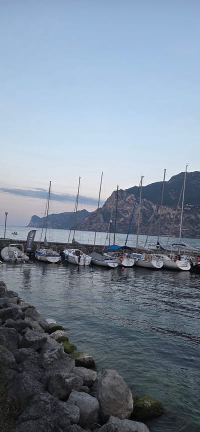 The image shows a marina in Torbole, Italy, with several sailboats docked along a stone pier. The boats are white with various colored sails and masts. In the background, mountains rise steeply from the water's edge, covered in vegetation. The sky is a pale blue with hints of pink, suggesting either sunrise or sunset. The water is calm, reflecting the boats and the sky. In the foreground, large rocks are piled along the shore. A lamppost is visible on the pier, and a banner with the text "WING & KITE" hangs from a pole. One small boat with two people is visible in the distance. The scene is serene and peaceful.