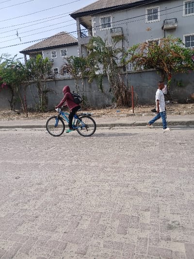 The image shows a street scene in Way, Nigeria, located at coordinates 6.429 latitude and 3.468 longitude. A person wearing a red hooded sweatshirt is riding a bicycle on a paved street in the direction of a two-story, gray building, while another person wearing a gray shirt and blue jeans walks away from the camera. There are power lines and trees in the background. The ground is covered in grey brick pavers, which are a common paving material in Nigeria. The scene depicts an ordinary day in Way, Nigeria.