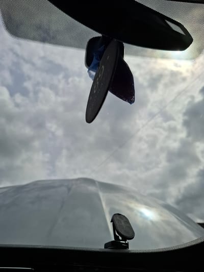 An exterior view from inside a vehicle shows a cloudy sky through the windshield. Hanging from the rearview mirror is a dark, oval-shaped object with some blue material attached. In the foreground, resting on the dashboard, is a black magnetic phone mount. The overall scene suggests a daytime environment, possibly in the city of Huntingdonshire, United Kingdom, though specific landmarks are not visible.