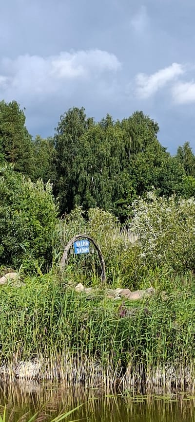 A blue sign with white lettering that reads "VALMA SADAM" is affixed to a wooden archway. The archway is situated amidst tall green reeds and other vegetation near the edge of a body of water. In the background, a dense line of trees forms the horizon. The sky above is overcast with dramatic clouds. The image appears to be taken on a day with mixed weather conditions. The location is identified as Valma, Estonia.