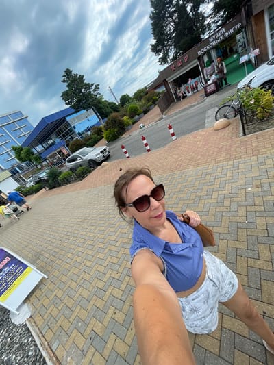 A woman is taking a selfie on a paved walkway in the city of Khostinskiy, Russia. She is wearing a blue striped crop top and white patterned shorts, with sunglasses and a brown purse. In the background, there is a building with "ТИР" and "МУЗЕЙ ВОСКОВЫХ ФИГУР" signs, which translates to "Shooting Range" and "Wax Figure Museum". There are also trees, a silver car, a bicycle, and other pedestrians in the scene. The sky is cloudy.