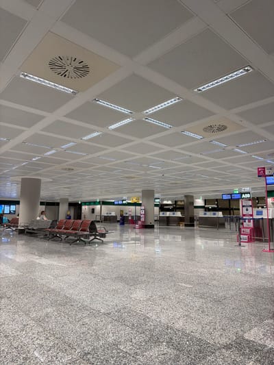 The image depicts a large, brightly lit indoor space, likely an airport terminal in Ferno, Italy, given the presence of airport-related signage and amenities. The ceiling is a grid of white panels, interspersed with recessed linear fluorescent lights and circular air vents. The floor is a highly polished, speckled gray granite.
In the foreground, rows of empty red and gray waiting chairs are arranged. To the left, a single person is seated in one of the gray chairs, looking towards a screen or display. Further into the background, a series of check-in counters or service desks are visible, some with electronic displays showing flight information. Several stanchions with signage, including one indicating "A59" and another with "WIZZ AIR," are positioned throughout the area. The overall environment suggests a quiet period, possibly early morning or late at night, with minimal activity. The polished surfaces reflect the overhead lighting and create a sense of spaciousness and order.