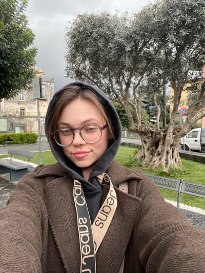 A young, light-skinned female subject is depicted in a selfie, centered in the frame, in a public green space in Scafati, Italy, under an overcast sky. She has light brown hair, wears circular-rimmed eyeglasses, and exhibits a slight smile. Her attire consists of a dark grey hooded sweatshirt with the hood up, worn under a dark brown overcoat with a discernible patterned texture. A beige and black strap featuring "Calvin Klein Jeans" text is visible across her chest.

The background shows an urban park setting. To the subject's immediate left, a light-colored, rectangular stone bench is present. Further left and behind, a multi-story building, possibly residential, displays weathered light-colored walls, dark windows, and a green railing on a lower balcony. A white sign, possibly indicating Wi-Fi, is mounted on a pole near the building. To the subject's right and behind, a large, mature olive tree with a prominent, gnarled, multi-trunked base and silvery-green foliage dominates the midground. Another tree with dense green leaves is visible on the far left. A dark metal bench with an ornate design is partially visible in the bottom right corner. Beyond the olive tree, a paved road is visible, with a white utility van parked near a red and blue circular street sign. The ground consists of grey paving stones and well-maintained green grass areas.