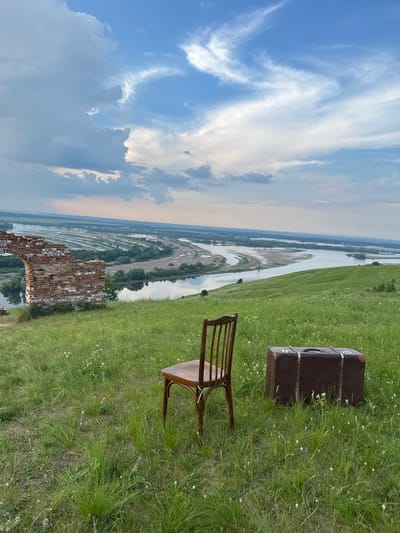 The image depicts a pastoral landscape with a winding river under a cloudy sky. In the foreground, a wooden chair and a vintage suitcase sit on a grassy hill. To the left, the ruins of a brick structure are visible. The background features a wide river meandering through a floodplain dotted with trees and distant settlements. The overall scene evokes a sense of tranquility and potential travel. The location is specified as Nizhniye Vyazovye, Russia.