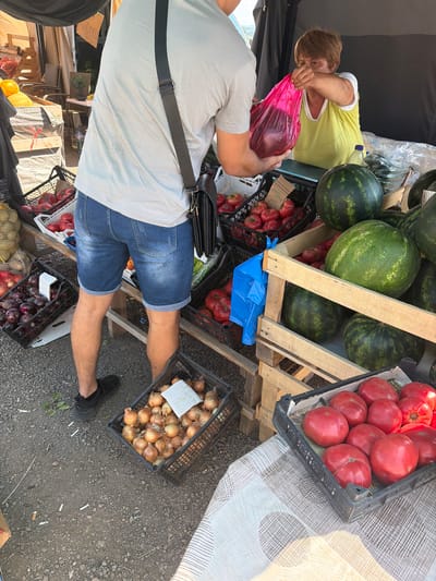 A man in blue denim shorts and a light grey t-shirt, wearing a black crossbody bag, is interacting with a woman at an outdoor market stall. The woman, wearing a yellow top over a white shirt, is handing a pink plastic bag to the man. The stall displays a variety of fresh produce including watermelons, tomatoes, and onions in crates and wooden boxes. The ground is covered with gravel. The background shows tents and other market structures, suggesting a bustling marketplace environment.
