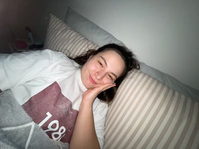 A person lies in bed with a striped pillow and blanket. They wear a white shirt with the number 1986 on it. Their hand is near their face as they smile into the camera. The room is dimly lit, showing a corner of the room with a glimpse of objects on the floor.