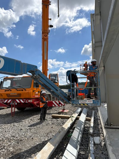 This image depicts a construction site in Jodłownik, Poland, under a bright, cloudy sky. In the foreground, a large orange Grove crane truck is parked on a gravel surface. A man wearing a high-visibility orange vest and a white hard hat stands near the truck. To the right of the truck, a blue aerial work platform (cherry picker) is elevated, carrying two construction workers. One worker, also in an orange vest and white hard hat, is actively working on a large metal beam attached to a building structure. Another worker in a yellow hard hat is visible in the platform behind him. The background shows more construction elements, including steel beams, and distant trees and hills. The scene suggests ongoing structural work, possibly on a new building or industrial facility.