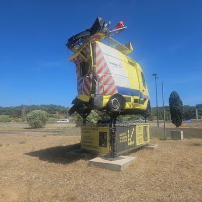 The image depicts a peculiar sculpture on a bright, sunny day with a clear blue sky. The sculpture is composed of a yellow and white striped van, seemingly damaged and elevated on a metal frame above a concrete base. The van has "VINCI AUTOROUTES" branding visible on its side. The top of the van is adorned with a complex metal structure, possibly salvaged car parts, including what appears to be a car bumper and a black grate. The van is tilted precariously, with one wheel visible.
The sculpture is situated in a dry, grassy area with scattered gravel. In the background, a highway with some traffic can be seen, bordered by a dense forest of green trees on a gentle slope. Two streetlights are also present near the sculpture.
A portion of the concrete base supports a yellow banner with black text and yellow circular graphics. The text on the banner reads, "RESPECTEZ LE CORRIDOR DE SÉCURITÉ" which translates to "RESPECT THE SAFETY CORRIDOR." Below this, it further states, "À l'approche d'un véhicule d'intervention: JE RALENTIS JE CHANGE DE VOIE," meaning "When approaching an intervention vehicle: I SLOW DOWN I CHANGE LANE." This suggests the sculpture is a public awareness display related to road safety.
While the specific location is Salles-d'Aude, France, there are no people visible in the image. The scene captures an outdoor environment characterized by arid vegetation and a highway backdrop, highlighting the striking art installation.