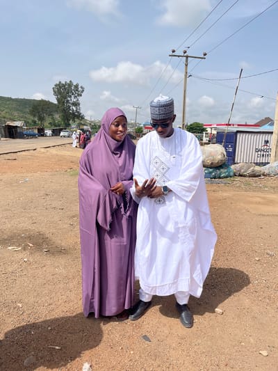 A man and woman are standing next to each other on a dirt road in Naraguta Hausawa, Nigeria. The man is wearing a white traditional outfit and a patterned hat, along with sunglasses. The woman is wearing a purple hijab. The background includes a road, a small hillside, and a few structures, along with utility poles and wires. In the distance is a container with "DANGOT CEMENT" printed on it.