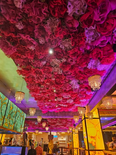 A vertically oriented, high-angle photograph showcases a decorative interior in Armação Dos Búzios, Brazil. Dominating the left side is a textured wall covered densely with fabric flowers in shades of pink, red, and light purple. Small, ornate chandeliers are suspended from the ceiling along the floral wall's edge.
To the right of the floral wall, the photograph captures a segment of the interior with a brightly lit, yellow-colored ceiling featuring a decorative pattern. Multiple chandeliers continue to be suspended from this ceiling as well.
A portion of the interior, including shelves stocked with items that may be merchandise, and people, can be seen in the background. In the foreground, there is a reflective, patterned surface in shades of green and gold.