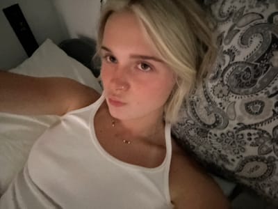 A young Caucasian woman with blonde hair and a nose ring lies in bed, looking at the camera. She is wearing a white ribbed tank top and a delicate necklace. The background shows a white pillow and a patterned duvet in shades of gray and white. The image appears to be a selfie taken in a bedroom. The provided crops offer closer views of her face, neck, and clothing. There are no discernible elements in the image that indicate the specific location of Squamish, Canada.