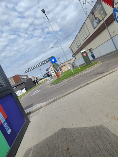 This image, captured in Arad, Romania, depicts an outdoor scene at what appears to be a commercial establishment, possibly a drive-thru service. In the foreground, a concrete surface transitions into a paved area with cobblestone patterns. To the left, a partially visible structure with a blue and purple panel featuring an image of a plastic bottle and recycling symbols suggests a waste or recycling bin.
Dominating the midground is a paved roadway designed for vehicles, indicated by white arrows painted on the surface. A large, striped barrier, possibly for traffic control, spans the roadway, displaying the number "2.70" and an upward-pointing blue arrow. Further down the road, several buildings are visible. To the right, a light-colored, large building with a brown-trimmed facade features a large advertisement for "G-SHOCK CENTRAL CEE" with an image of a person. A security camera is mounted on this building. Adjacent to this building is a dark green pole with a red and white circular sign, indicating a prohibition. A person in a blue uniform is visible near the entrance of the building.
In the background, other buildings are present, including a brown brick structure and several residential-style buildings with white and beige facades. The sky above is filled with white, fluffy clouds against a light blue backdrop. Streetlights are positioned along the roadway. The overall scene suggests an active commercial area with infrastructure for customer traffic.