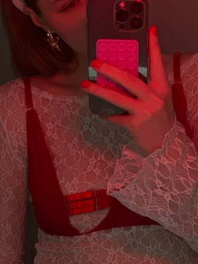 A woman's perspective is captured in a selfie. The image is lit with red light. Her left hand holds a smartphone with a glittery grey case and a pink, circular-patterned grip. She's wearing a white lace garment over a red piece of clothing. Her hair is reddish-brown and falls over her left shoulder, displaying a gold and red earring. The focus is on the hand, phone, and the lace material.