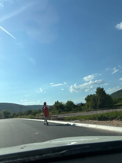 A person is riding an electric scooter on a paved road in Lastva Grbaljska, Montenegro. The person is wearing a red backpack and is facing away from the camera. The road is curved and appears to be under construction, with piles of dirt and debris on the side. There are hills and trees in the background, and the sky is blue with some clouds. The image is taken from inside a car, with the dashboard visible in the foreground.