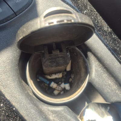 The image displays an open ashtray filled with cigarette butts and ash. One cigarette butt is clearly visible with the brand name "Winston" printed on it. The ashtray is located within a car's center console. The surrounding environment appears to be dusty, with specks of light reflecting off surfaces. The ashtray lid is open, exposing its contents. The overall scene is consistent with a used ashtray in a vehicle.