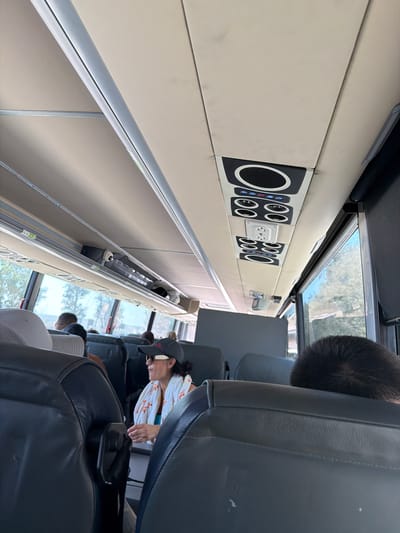 The image shows the interior of a bus or train. The camera is positioned looking down the aisle, with rows of seats visible on the right. Passengers are seated in some of the seats. A woman in a white and red patterned shirt and a gray cap is visible. The seat in the immediate foreground is dark gray. The left side of the image shows the interior wall of the vehicle, with a series of black panels with speaker-like grills and controls, including power outlets. Above the panels, a window offers a glimpse of trees and sky outside. The location is uncertain, there is no information available for the Fontana, United States of America region.