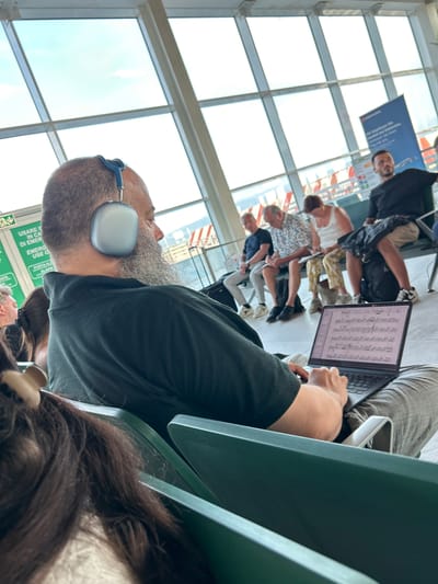 In a well-lit, modern waiting area, likely an airport or train station, a group of people are seated, some engaged with electronic devices. In the foreground, a man with a beard and headphones is facing away from the camera, engrossed in a laptop displaying what appears to be musical notation. To his left, other individuals are also seated, including a woman in a white top and a man in a patterned shirt. Behind them, a large window overlooks an outdoor scene, possibly indicating daytime with bright, natural light. A blue banner with text, partially visible and featuring the "edelweiss" logo, suggests a travel or airline-related context, potentially an airline's services or a travel agency. The overall atmosphere seems relaxed, with individuals occupied with their personal activities while waiting. Visible text includes "USARE IN CA DI EMER EMERG USE O PORTA AL ALARM" and "edelweiss Wie beartassen She avalih tech Edelweiss. Comassen Sie liven". The presence of travel-related signage and the diverse group of people suggests a travel hub.