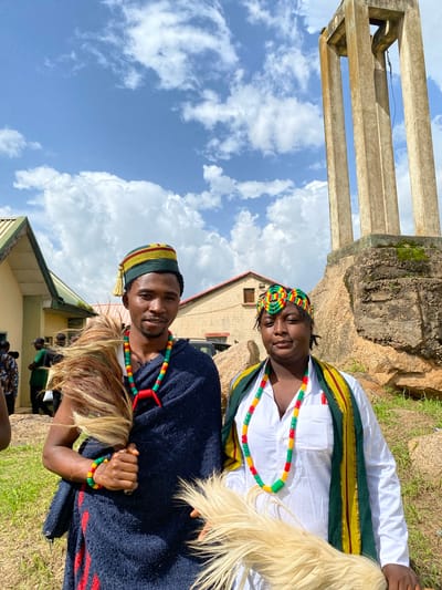The image depicts two individuals adorned in traditional attire, likely participating in a cultural event or ceremony in Wang, Nigeria. The subjects are a man and a woman, both wearing colorful beaded necklaces and bracelets in red, yellow, and green. The woman is wearing a white shirt with a green and yellow sash draped across her shoulders. The man is wearing a dark blue garment over a white shirt and a cap with similar colorful accents.
The setting appears to be outdoors, with a backdrop of a rocky hill and a cloudy blue sky. In the foreground, lush green grass is visible. A concrete structure, possibly part of an ancient monument or building, stands to the left. The sunlight suggests it is daytime, and the clouds indicate it might be a partly cloudy day. The overall atmosphere seems festive and celebratory, with the subjects looking relaxed and engaged.
The image is oriented vertically. The people are positioned in the center, with the natural landscape and structures surrounding them. The bright colors of their clothing and accessories stand out against the more muted tones of the environment. The expressions on their faces suggest a sense of pride and enjoyment. No visible text is present in the image.