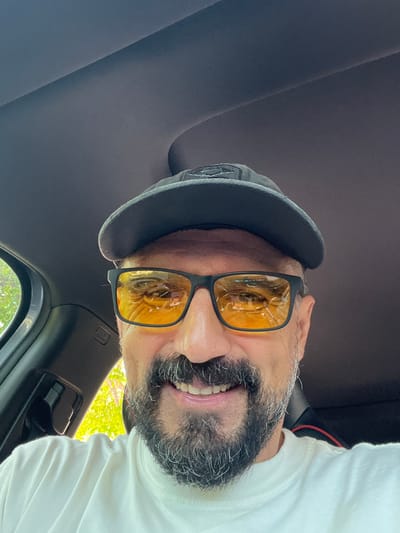A man wearing a black baseball cap and yellow-tinted sunglasses is taking a selfie inside a car. The car's interior is visible, with the dark headliner of the car above and a portion of the driver's side door and dashboard to the left. Through the car window, bright green foliage and trees are visible, suggesting an outdoor setting. The man has a beard and appears to be smiling. The image is framed from the chest up. The location is not specified in the image.