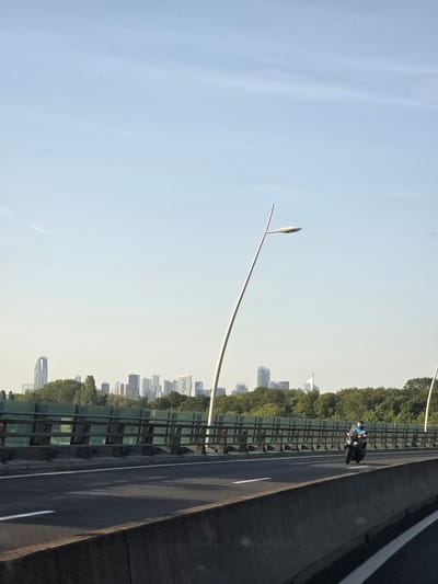 The image depicts a view from a highway overpass in Boulogne-Billancourt, France, looking towards a distant city skyline. In the foreground, a dark concrete barrier lines the side of the road, with a grey asphalt surface stretching into the distance. A white line is visible on the road, marking a lane divider. A motorcycle, ridden by a person wearing a blue jacket and helmet, is traveling on the highway from left to right. Behind the motorcycle, a green metal fence with horizontal wooden beams runs along the edge of the overpass, separating it from a line of lush green trees. Beyond the trees, a cluster of modern skyscrapers, indicative of a city center, is visible against a pale blue sky. A tall, slender lamppost with a curved arm and a light fixture stands on the right side of the highway. The overall scene is bathed in bright daylight, suggesting a clear, sunny day.