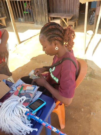 In Kwamba, Nigeria, a young woman with braided auburn hair is counting money, holding a wad of cash in her hands. She is wearing a maroon t-shirt with green lettering that appears to read "CHANGE MAN" and a sparkling earring in her left ear. She is seated on a blue plastic chair, with an orange chair visible behind her. On the blue chair, a mop with a floral-patterned handle, a stack of books or notebooks, and a small blue electronic device are placed. The scene appears to be outdoors, with a dirt ground and sunlight casting shadows, suggesting it is daytime. The background shows some wooden structures, possibly a market stall or dwelling, and a metal grate. The woman's expression is one of concentration as she handles the money.
