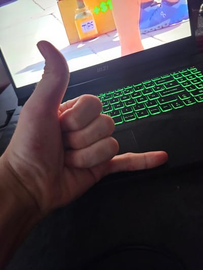 The image shows a person's hand making a "shaka" sign in front of a laptop with a green-lit keyboard. The laptop screen displays a scene that appears to be from a video game or animation, featuring a bottle labeled "TIPS" and a dollar sign. The background on the screen shows a floor that looks like cobblestones or tiles and a person's lower legs wearing blue athletic shoes. The scene is indoors, as indicated by the laptop and keyboard. There is no identifiable information in the image to suggest a location in the city of Versailles, France.