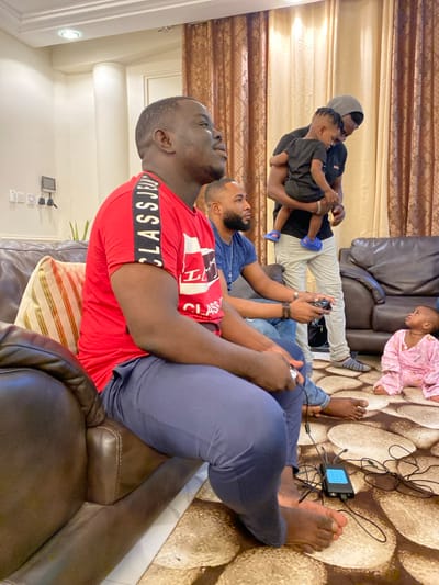 The image, rotated 90 degrees clockwise, captures a casual indoor scene, likely a living room, in Adenta Municipality, Ghana. Several individuals, primarily males, are present.
In the foreground, a man wearing a red t-shirt with "CLASS JEANS" printed on it is seated on a brown sofa, engaged in playing a video game with a controller in his hands. Beside him, another man in a blue t-shirt is also seated and holding a gaming controller, looking towards the right, presumably at a screen beyond the frame. Both men are barefoot.
In the mid-ground, a man wearing a grey hoodie or cap is holding a young child dressed in a black top. They appear to be observing the gaming activity. On the patterned carpet to the right, a baby or toddler in a pink patterned sleepsuit is sitting, also looking towards the right.
On the floor, between the gamers and the baby, a small electronic device, likely a gaming console or mini-PC, is visible with multiple connecting wires. The room is furnished with brown leather-like sofas and features patterned gold and brown curtains in the background. Indoor lighting fixtures are visible on the ceiling. The scene depicts a relaxed gathering, possibly a family enjoying leisure time together with video games.