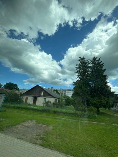 The image captures a view from a moving vehicle on a sunny day in Olaine, Latvia. A rural or suburban landscape is depicted, dominated by a clear blue sky filled with large, fluffy white clouds. In the foreground, a paved walkway or road edge is visible on the left. The middle ground features a green grassy lawn and a quaint, old brick house with a dark wooden roof. A tall, dense evergreen tree stands to the right of the house, obscuring a portion of a multi-story apartment building in the background. The overall impression is one of a tranquil summer day in a residential area.