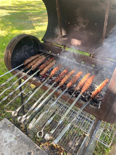 In the city of Znamenskoye, Belarus, a close-up shot captures a grilling process on a sunny day. The focus is on a rustic, barrel-shaped grill, its metal exterior weathered and stained, suggesting frequent use. Inside, glowing charcoal embers provide heat for several skewers of seasoned meat, likely lula kebab, arranged in two rows. The meat is browned and appears to be in the final stages of cooking, with visible smoke rising from the grill. The skewers themselves are made of metal, with twisted handles for easy rotation. They rest on the grill's metal racks. Surrounding the grill is a patch of green grass, some fallen leaves, and a concrete slab in the foreground, indicating an outdoor setting, possibly a backyard or park area. The overall impression is one of a leisurely outdoor meal being prepared.