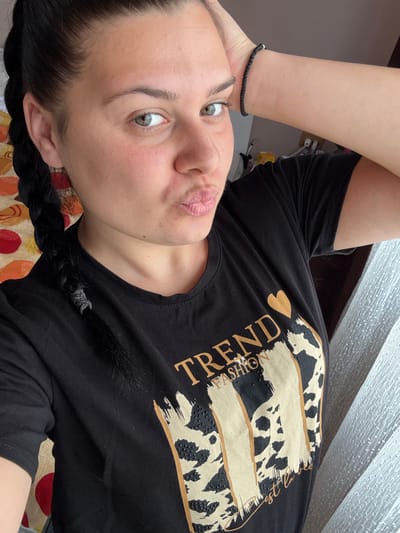A young woman with light green eyes, dark hair in a braid, and fair skin poses for a selfie. She is wearing a black t-shirt with a design on it, and a black braided bracelet on her wrist. Her lips are pursed, and her right hand is resting on her head. The background includes a curtain and a patterned surface. The location is not discernible from the image.