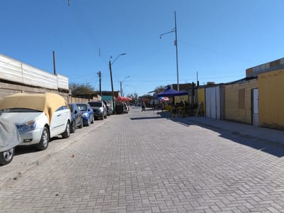 This image depicts a street scene in Pozo Almonte, Chile. The street is paved with interlocking bricks, and on the left side, several cars are parked along the sidewalk. One car is partially covered with a yellow tarp. The buildings lining the street appear to be simple structures, some with corrugated metal siding and others with yellow or beige painted facades.
In the distance, there are more parked cars and a few people walking along the sidewalk. On the right side of the street, there are some market stalls with blue and red umbrellas providing shade. Several people are gathered at these stalls, suggesting a commercial or social gathering. The sky is clear and blue, indicating a sunny day. The presence of utility poles and wires suggests an urban or semi-urban environment. There is no visible text in the image. The overall impression is that of a typical daytime scene in a small town or city.