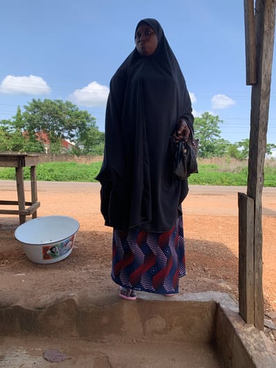 A woman stands outdoors wearing a black hijab and a patterned skirt. She holds a black bag in her right hand. In front of her, to her left, is a white oval basin with colorful images on its side. The ground is covered in reddish-brown dirt. Behind the woman, a road is visible, bordered by green vegetation and trees. In the background, a building with a red roof can be seen. The scene appears to be in Gbeleko, Nigeria, based on the context provided. The sky is blue with scattered white clouds. The woman is standing on a raised concrete platform. A wooden post is visible to the right of the frame.