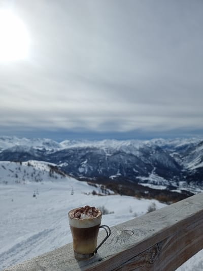 The scene depicts a snow-covered mountain landscape at a ski resort in Cesana Torinese, Italy, viewed from an elevated vantage point. In the immediate foreground, a clear glass mug containing a layered beverage, topped with white whipped cream and dark powder, rests on a weathered wooden railing. The mug features a metal holder with a handle. The midground consists of extensive snow-covered ski slopes exhibiting visible tracks and a ski lift infrastructure with poles and cables. Scattered trees are present on the slopes. The background is composed of distant, snow-capped mountain ranges under a partially cloudy sky. A bright sun, manifesting as a glare, is visible in the upper-left quadrant of the sky. No human figures or active interactions are observed.
