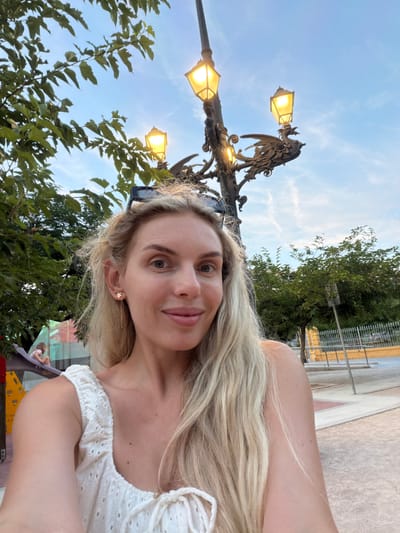 A medium shot captures a blonde woman with fair skin and light blue eyes taking a selfie outdoors. She is smiling slightly and looking directly at the camera. She has blonde hair that falls past her shoulders, and is wearing a white top with eyelet detailing. A gold earring in the shape of a star with a dangling chain is visible in her left ear. Behind her, a streetlamp with three illuminated yellow lights and an ornate, dragon-like fixture is visible against a blue sky with wispy white clouds. To the left, lush green tree branches with large leaves partially obscure the sky. In the background, a playground structure with a red and yellow slide and a young child on the slide can be seen. Further back, trees and a metal fence are present, suggesting an urban park or public space. The woman's location is identified as Benimàmet-beniferri, Spain.