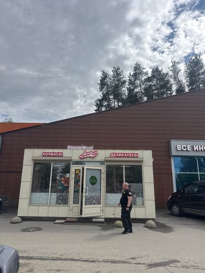 A man in a black uniform, possibly a security guard, stands outside a building with a brown tiled facade. The building appears to be a store with large glass windows displaying various products and decorations, including images of food items. Two signs are visible above the entrance, one in red and white lettering that reads "КОЛБАСЫ" and another in white lettering that says "ДЕЛИКАТЕСЫ". Above these, a larger sign in white letters on a gray background reads "ВСЕ ИНО". The man is facing to the right, with his hands behind his back. A black car is parked nearby, and large rocks are placed on the pavement, possibly as barriers. The sky is cloudy, indicating overcast weather. The scene is likely taking place outdoors in a commercial or retail area in Kuzmolovskiy, Russia. The overall mood appears neutral, with no obvious emotional expressions.