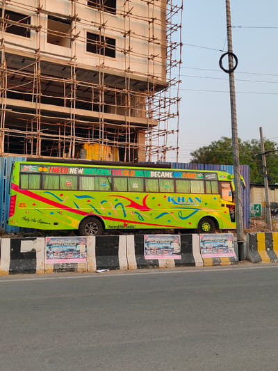The image displays a static street scene in Anantapur, India, under clear daylight conditions. A prominent lime-green bus, featuring vibrant pink, blue, and yellow abstract designs, is parked in the midground. Text on the bus includes "I NEED NEW HATERS THE OLD ONES BECAME MY FANS" along the top band, "KHAN HOLIDAYS A KHAN HOLIDAYS GROUP OF CONCERN" on the side, and "Incredible India!" and "Fully Air Suspension" near the bottom. Its registration number, "AP39 TB.1115", is visible on the rear left, and "MERE PRABHU MERI ANANTAPUR" is partially legible near the front.

To the left of the bus, a multi-story building under construction is visible, extensively covered by wooden pole and plank scaffolding. The building's concrete structure and numerous window openings are apparent. To the right of the bus, a tall, grey concrete utility pole stands, with black cables coiled and extending from it. Behind the bus, a blue corrugated metal fence separates the bus from green foliage and undeveloped ground.

In the foreground, a concrete road barrier with alternating black and yellow stripes spans horizontally. Three posters, primarily in Telugu script, containing images and advertisements, including the word for Anantapur, are affixed to this barrier. An asphalt road surface is visible in the lower portion of the image, in front of the barrier. No individuals are present in the scene.