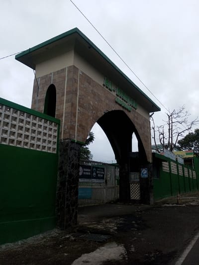 The image depicts an exterior view of an institutional entrance located in Kodarapalli, India, under an overcast sky. The primary subject is a large archway entrance, constructed with a light brown, rough-cut stone veneer on its pillars and arch, topped by a light beige upper section and a dark green flat roof or eave. Green block lettering spelling "AL-DAWAH" is affixed to the beige section of the archway, with smaller, less legible text beneath it.
To the left of the archway, a continuous dark green wall features a rectangular section composed of white, patterned, open-block screening. The base of the archway's pillars is clad in dark, irregular stone. Directly under the archway, a metal gate, appearing closed, provides the entrance. Attached to the left side of this gate are multiple signs, including a white sign with black text, partially legible as "DILARANG MEROKOK" (No Smoking), "BUANG SAMPAH PADA TEMPATNYA" (Dispose of trash in its place), and "BUKAN TEMPAT BERMAIN" (Not a playground). A blue sign with white text and a symbol, partially legible as "WISATA MIN," is affixed to the right pillar of the archway.
The ground in the immediate foreground consists of dark asphalt or compacted dirt with lighter, irregular patches. A metal drainage grate is embedded in the ground to the left. The green wall extends to the right of the archway, displaying recessed rectangular sections. Behind this wall, a tall, bare deciduous tree with numerous branches stands against the sky. Further in the background, another building with a green roof is partially visible. A thin, dark utility wire traverses the upper portion of the frame diagonally from top-left to mid-right. No individuals or specific activities are present in the scene.