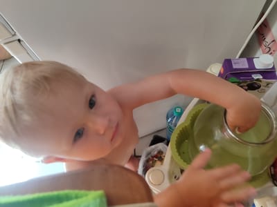 In this image, a toddler with blonde hair and blue eyes is the central subject. They are positioned at the top of the frame, looking down towards the camera. The toddler is partially undressed, suggesting a warm environment or a playful moment. Their hands are reaching out, one towards a clear glass bowl with a green silicone holder, and the other seemingly interacting with something out of frame.
The setting appears to be indoors, possibly a kitchen or a play area, given the presence of household items. Visible objects include a purple juice box with a white cap, a blue water bottle with a green label, and a clear plastic bag filled with colorful small objects, likely snacks or toys. A bright green object, possibly a mat or a toy, is in the foreground.
The lighting suggests it could be daytime, with a bright light source coming from the left, casting some shadows and highlighting the toddler's features. The overall mood seems playful and curious, as indicated by the toddler's direct gaze and active engagement with their surroundings.
The juice box has text on it, but it is mostly illegible. A pink banner with what appears to be Japanese text, "sushi," is visible on the right side of the frame.
The context indicates the location is Tuapse, Russia.