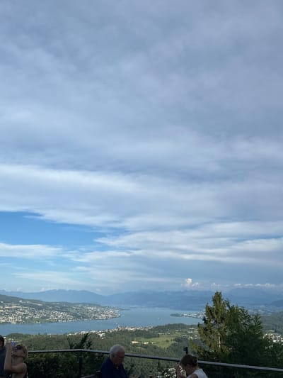 The image captures a panoramic view from a high vantage point overlooking a city nestled along a large body of water, with mountains in the distance. The sky is a mixture of blue and white clouds, with some areas appearing brighter than others. In the foreground, several people are present, seemingly seated or standing near a railing. To the left, a woman with blonde hair and sunglasses is looking upwards, holding a phone in her hand. Next to her, a man in a dark shirt is seated. In the mid-ground, a lush green landscape with scattered trees leads down to the city. The city itself is a densely populated area with many buildings of varying sizes. The water, likely a lake, is a deep blue and appears calm, with a few boats visible on its surface. The distant mountains form a hazy silhouette against the horizon. This scene is characteristic of a view from the area around Stallikon, Switzerland, likely from a restaurant or viewpoint offering vistas of Lake Zurich and the surrounding Alps.