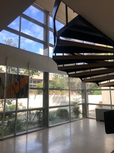 The image shows an interior space with a large window on the left side and a spiral staircase on the right side. The window provides a view of an exterior courtyard with a small garden and a white wall with windows. The spiral staircase is made of metal and wood, with a black railing and wooden steps. The ceiling of the space is white, and the floor is made of light-colored tiles. The image was taken at the location of the Agia Paraskevi Cultural Center in Agia Paraskevi, Greece, at the coordinates 37.999, 23.82. The scene is empty of people and appears to be quiet and peaceful. The image is taken from a low angle, giving the impression of looking up at the staircase.