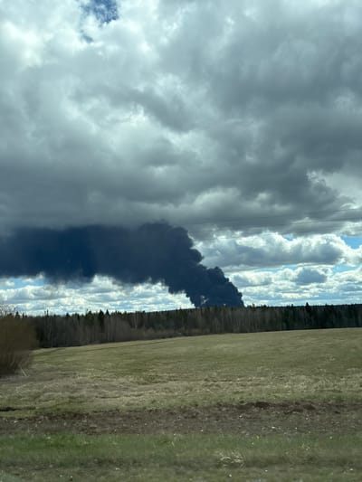 The image, when correctly oriented horizontally, captures a rural landscape dominated by a large plume of dark smoke rising into the sky. In the foreground, a field of dry, mixed green and brown grass stretches across the lower portion of the frame. Faint tracks or variations in the terrain are visible in the field.
A dense treeline of dark evergreen trees forms the middle ground, creating a distinct horizon line. Directly behind this treeline, towards the center-left, a massive column of black, billowing smoke ascends skyward, indicating a significant fire. The smoke is dark and heavy, partially obscuring the sky above.
The sky itself presents a dramatic backdrop. Heavy, dark grey clouds cover much of the upper left and center, suggesting overcast conditions or an approaching storm. However, patches of bright blue sky with fluffy white cumulus clouds are visible towards the upper right and in gaps between the darker clouds, providing a contrast in lighting. Several power lines run horizontally across the upper-middle section of the image, passing above the smoke and treeline, suggesting nearby infrastructure.
The scene depicts an event of active smoke production from a fire during daytime hours, under mixed weather conditions. No individuals or specific sources of the fire are visible. The setting appears to be a natural, open area. This event is contextualized as occurring near Башкултаево, Russia.