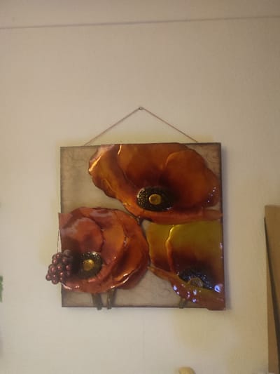 The image displays a three-dimensional, stylized artwork depicting three poppy-like flowers on a textured, light gray rectangular panel, prominently mounted on a plain, light-colored wall in Southampton, United Kingdom. The artwork features a glossy finish, suggesting materials like glazed ceramic, resin, or painted metal.
The flowers are predominantly in shades of orange, red, and yellow. The largest flower, located at the top-right, exhibits orange-red petals and a dark center with a golden-yellow accent. A second, medium-sized flower is positioned below it on the left, displaying similar red-orange petals and a dark center with a golden accent. Attached to the left side of this second flower is a small cluster of dark red or purple spherical objects, resembling berries. A third, smaller flower is partially visible at the bottom-right, showing orange-yellow petals and a dark center.
The artwork panel is suspended by a thin, reddish string or cord attached to a single metallic hook or nail in the wall, visible just above the panel's top edge. The wall itself is off-white or light cream. Partial glimpses of other objects are present: a green item on the far left, a vertical brown object on the far right, and a small, light-toned, indistinct object at the bottom center, resting against the wall. No individuals, actions, or direct interactions are visible within the frame.