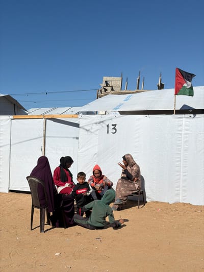 This image, captured in Jabalia, Palestinian Territory, depicts a group of six individuals on sandy ground outside temporary shelters under a clear blue sky.
The environment consists of light brown sand and dirt, with scattered small debris. Several white temporary shelters, constructed from tarp-like material over wooden frames, dominate the midground. One prominent shelter features the black numeral "13" centrally on its side. Faint blue "UNICEF" and "UNRWA" logos are visible on the tarp roofs of some shelters. A Palestinian flag, displaying red, black, white, and green, flies from a pole attached to the top right of a shelter. In the background, behind the shelters, partially constructed concrete buildings with exposed rebar are visible. Overhead, thin black electrical wires traverse the scene.
Six individuals are present, comprising four adult females and two children, engaged in an informal gathering.
1. An adult female, dressed in a light brown patterned dress and head covering, sits on a dark chair on the right, gesturing with her right hand while facing the group.
2. An adult female, wearing a red top and black head covering, sits on a chair to the left of the first woman, looking downwards.
3. An adult female, clad in a dark purple garment and head covering, sits on a black chair with her back partially to the camera, facing the group.
4. An adult female, wearing an orange hooded top, sits directly on the ground, facing the group, holding a small, light-colored object in her hands.
5. A young boy, wearing a black hoodie with "Noytime" in red text, sits on the ground between the woman in red and the woman in orange, looking downwards at an unseen object.
6. A child, wearing a green hooded top and dark pants, sits on the ground with their back partially to the camera, appearing to interact with the dirt surface.