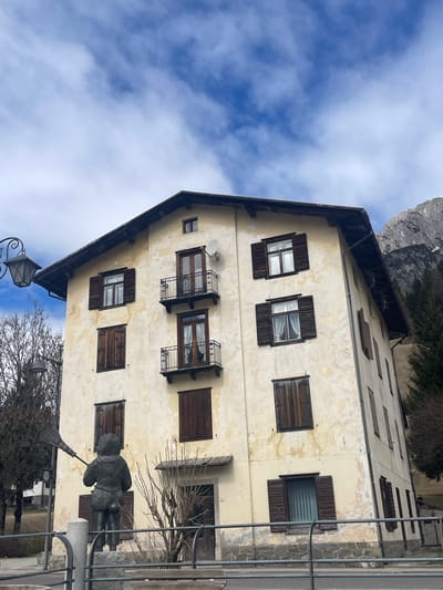 The image captures a tall, narrow building in Sappada / Plodn / Sapade, Italy. The structure features multiple stories, each adorned with rectangular windows framed by dark brown wooden shutters. Some of the shutters are open, revealing glimpses of the interiors. The building's facade is painted in a light yellow hue, exhibiting patches of peeling paint, adding a sense of age and wear to the structure. The roofline is dark and sharply angled. A small satellite dish is mounted on the building's side.
In the foreground, a metal sculpture of a man holding a tool is visible. A wrought-iron fence runs along the front of the scene. Bare trees and shrubs are scattered nearby, suggesting a winter or early spring season. The sky above is bright blue, partially covered with wispy white clouds. In the background, a rocky mountain face can be seen, hinting at the mountainous terrain of the area.