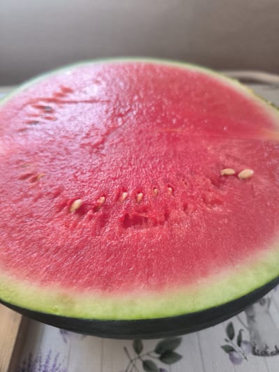 The image shows a close-up, slightly angled view of a cut watermelon, displaying its vibrant pink flesh and scattered dark seeds. The rind is visible, showing a transition from the pale green inner layer to the darker green outer peel. The watermelon is placed on a surface covered with a light-colored fabric featuring a floral pattern, with hints of purple flowers. The lighting suggests it's daytime, possibly indoors with natural light. The scene is simple and focused on the fruit, with no other objects or people prominently visible. The texture of the watermelon's flesh, with its slightly glistening appearance, is a notable detail. No text is visible in the image.