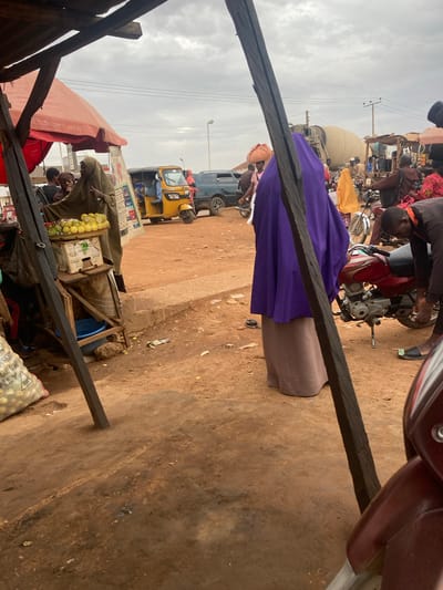 The image depicts a busy outdoor market scene in what appears to be a city in Nigeria, possibly in the vicinity of phone number 0801712591. The ground is primarily dirt and appears well-trodden. In the foreground, a wooden structure partially frames the view, likely a vendor's stall. A person in a purple garment is centered in the middle ground. Various other individuals, motorcycles, and a yellow three-wheeled vehicle (a "keke") are visible further back. Merchandise, including what appears to be fruits, are visible at vendor stalls. The sky is overcast.