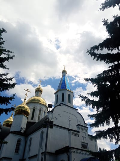 The image shows a low-angle view of the Church of the Intercession of the Most Holy Theotokos, located in Yessentukskaya, Russia. The church features multiple golden domes topped with Orthodox crosses and a distinctive blue-tiled spire. The building is predominantly white with architectural details and decorative elements. Dark green pine branches frame the scene from the left and right, creating a sense of being enveloped by nature. The sky above is a mix of white clouds and patches of blue, suggesting a partly cloudy day. The lighting indicates it is daytime. There are no people visible in the image.
